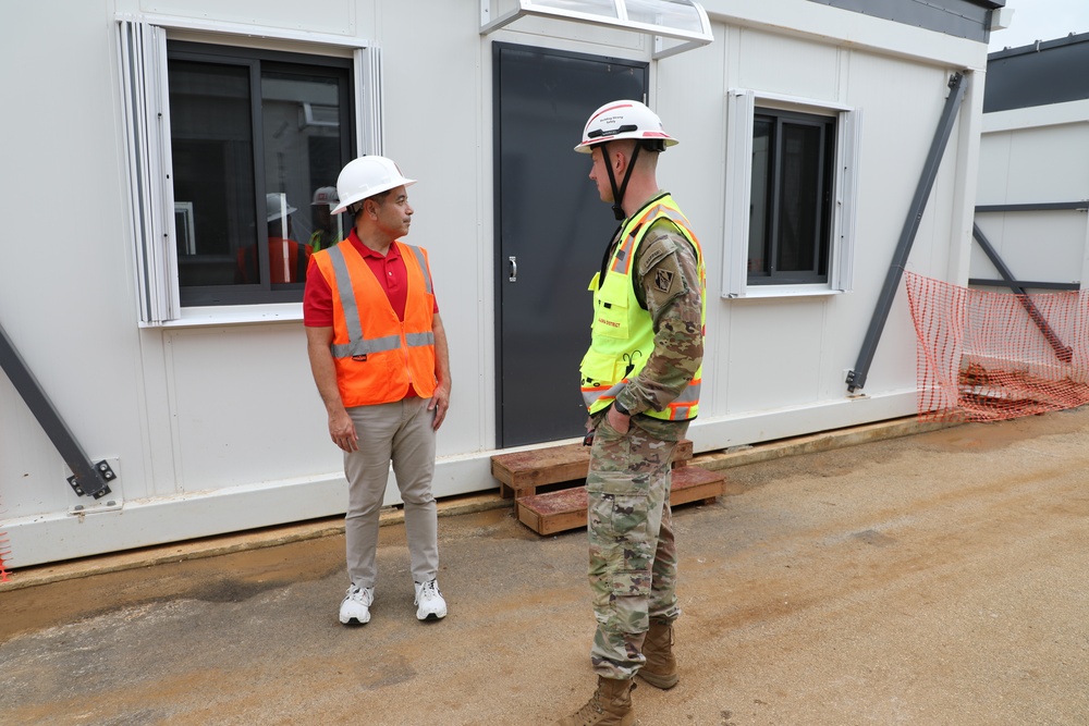 Two individuals are conversing outside a modular or prefabricated building at what appears to be a construction or military installation site. One person is wearing civilian attire with an orange high-visibility vest and white hard hat, while the other is in military camouflage (OCP/MultiCam) uniform with a yellow high-visibility vest and white hard hat. The setting includes safety fencing and construction materials, suggesting an active work zone.