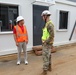 Two individuals are conversing outside a modular or prefabricated building at what appears to be a construction or military installation site. One person is wearing civilian attire with an orange high-visibility vest and white hard hat, while the other is in military camouflage (OCP/MultiCam) uniform with a yellow high-visibility vest and white hard hat. The setting includes safety fencing and construction materials, suggesting an active work zone.