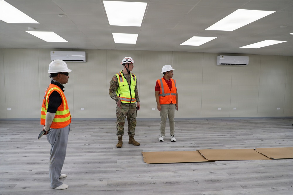 Three individuals are standing inside a newly completed or nearly finished interior space, likely during a construction inspection or walkthrough. One person is wearing military camouflage uniform with a safety vest, while the other two wear orange high-visibility vests and white hard hats. The room features installed LED panel lighting, wall-mounted air conditioning units, and laminate flooring.