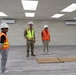 Three individuals are standing inside a newly completed or nearly finished interior space, likely during a construction inspection or walkthrough. One person is wearing military camouflage uniform with a safety vest, while the other two wear orange high-visibility vests and white hard hats. The room features installed LED panel lighting, wall-mounted air conditioning units, and laminate flooring.