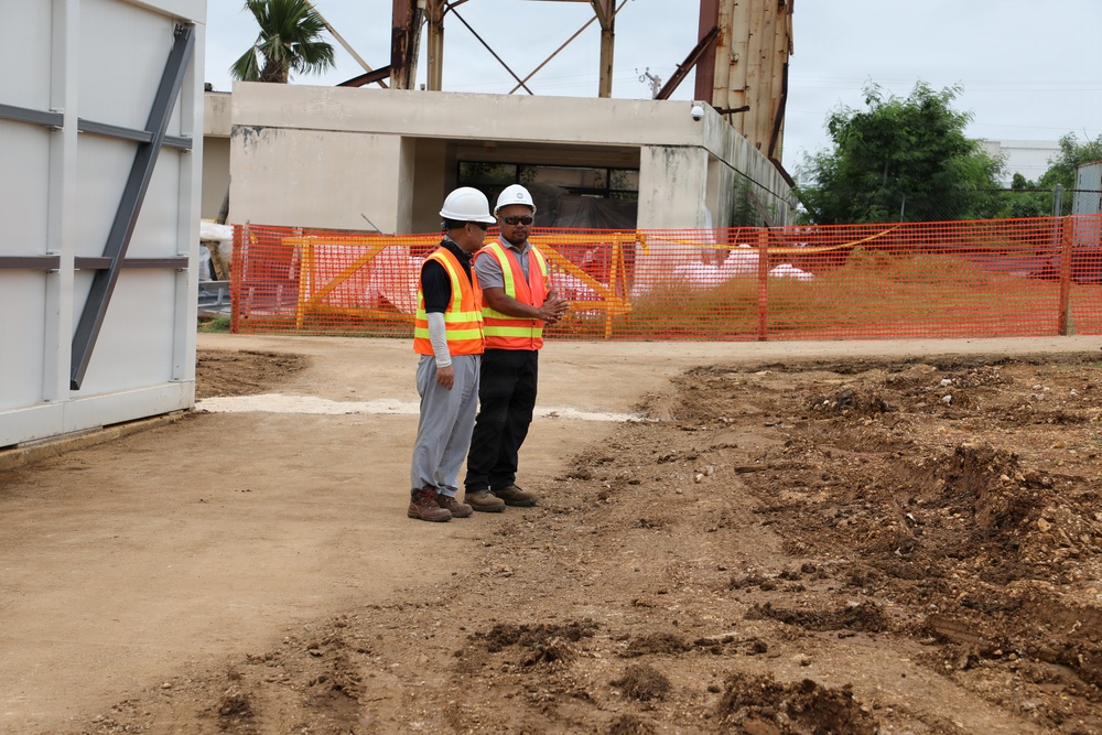 Two construction workers wearing orange high-visibility vests and white hard hats are conversing at an active construction site. The site shows significant ground disturbance with exposed soil, excavated areas, and orange safety fencing in the background. A concrete structure and metal framework are visible behind them.