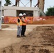 Two construction workers wearing orange high-visibility vests and white hard hats are conversing at an active construction site. The site shows significant ground disturbance with exposed soil, excavated areas, and orange safety fencing in the background. A concrete structure and metal framework are visible behind them.