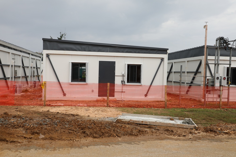 The image shows a construction or installation site featuring modular/prefabricated white buildings with dark roofing. The structures appear to be newly erected and are surrounded by orange safety fencing, indicating active construction. The foreground shows disturbed earth and a concrete pad, suggesting ongoing site development.