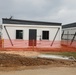 The image shows a construction or installation site featuring modular/prefabricated white buildings with dark roofing. The structures appear to be newly erected and are surrounded by orange safety fencing, indicating active construction. The foreground shows disturbed earth and a concrete pad, suggesting ongoing site development.