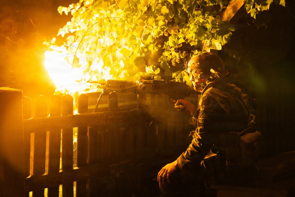 A heavily equipped soldier in full tactical gear is firing a weapon at night, producing a large muzzle flash that illuminates the surrounding vegetation and wooden fence. The scene depicts a nighttime combat or training operation in what appears to be a residential or rural area. The intense orange light from the muzzle blast dominates the composition.