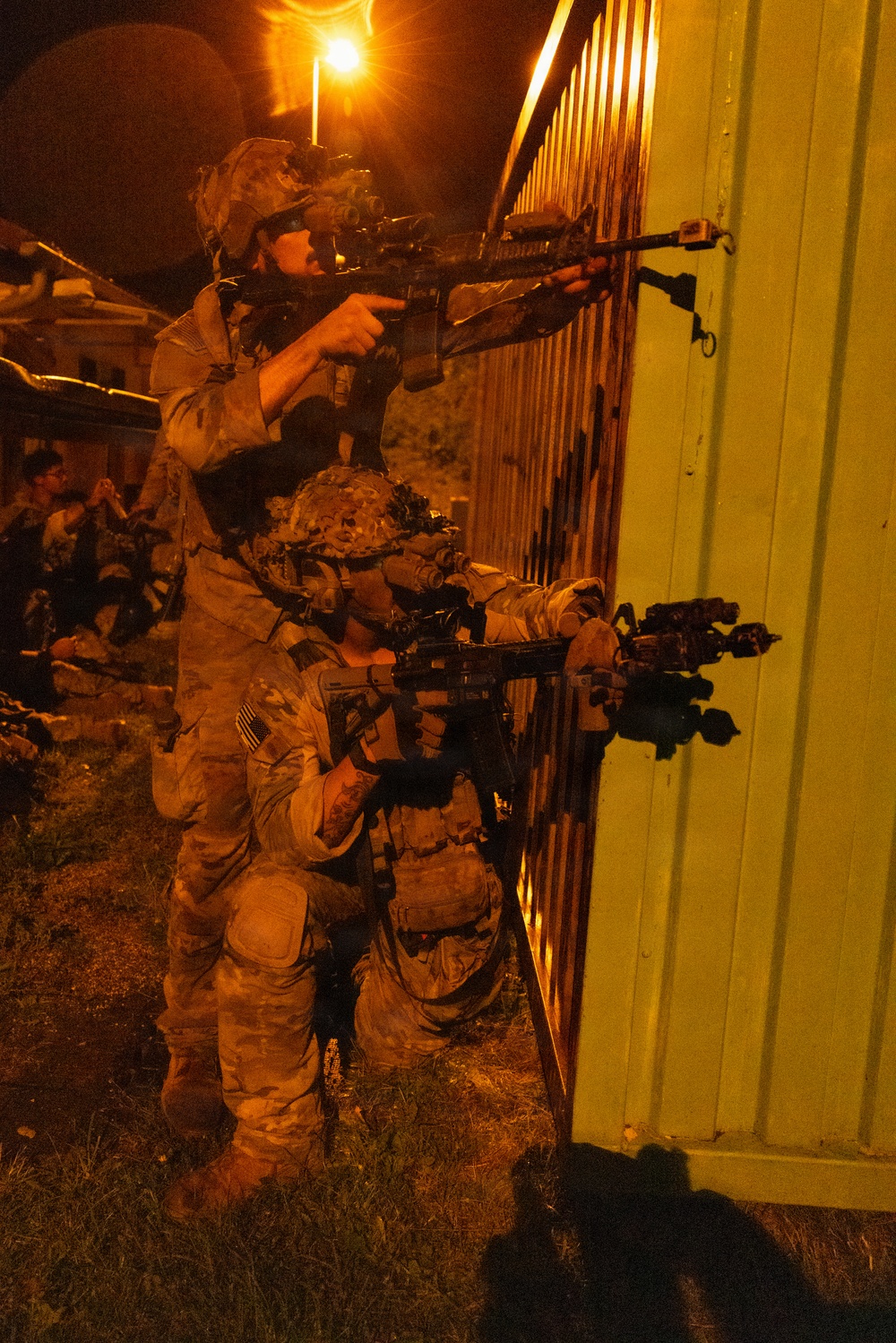 Two armed soldiers in full combat gear are positioned against a corrugated metal structure during a nighttime operation. One soldier stands while aiming a rifle over the top, while a second soldier kneels below aiming outward around the corner. Additional personnel are visible in the background, suggesting a larger unit operation or training exercise.