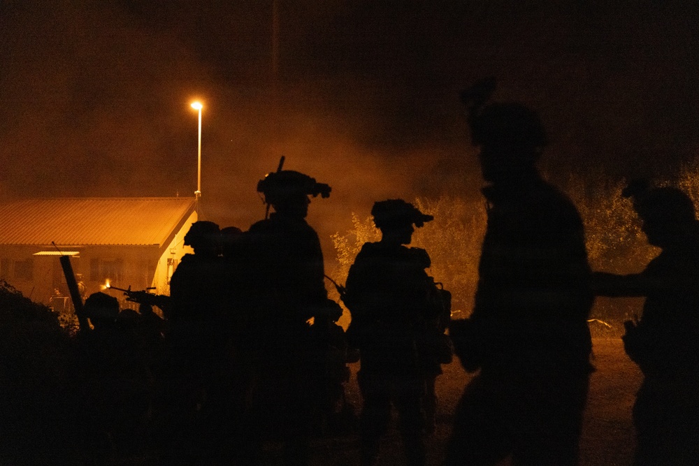 A nighttime scene showing multiple silhouetted figures in full tactical/military gear, including helmets with mounted equipment, moving in a group. The scene is illuminated by warm orange ambient light from a street lamp and what appears to be a structure in the background. Smoke or haze is visible in the atmosphere, adding to the dramatic low-light conditions.