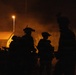 A nighttime scene showing multiple silhouetted figures in full tactical/military gear, including helmets with mounted equipment, moving in a group. The scene is illuminated by warm orange ambient light from a street lamp and what appears to be a structure in the background. Smoke or haze is visible in the atmosphere, adding to the dramatic low-light conditions.