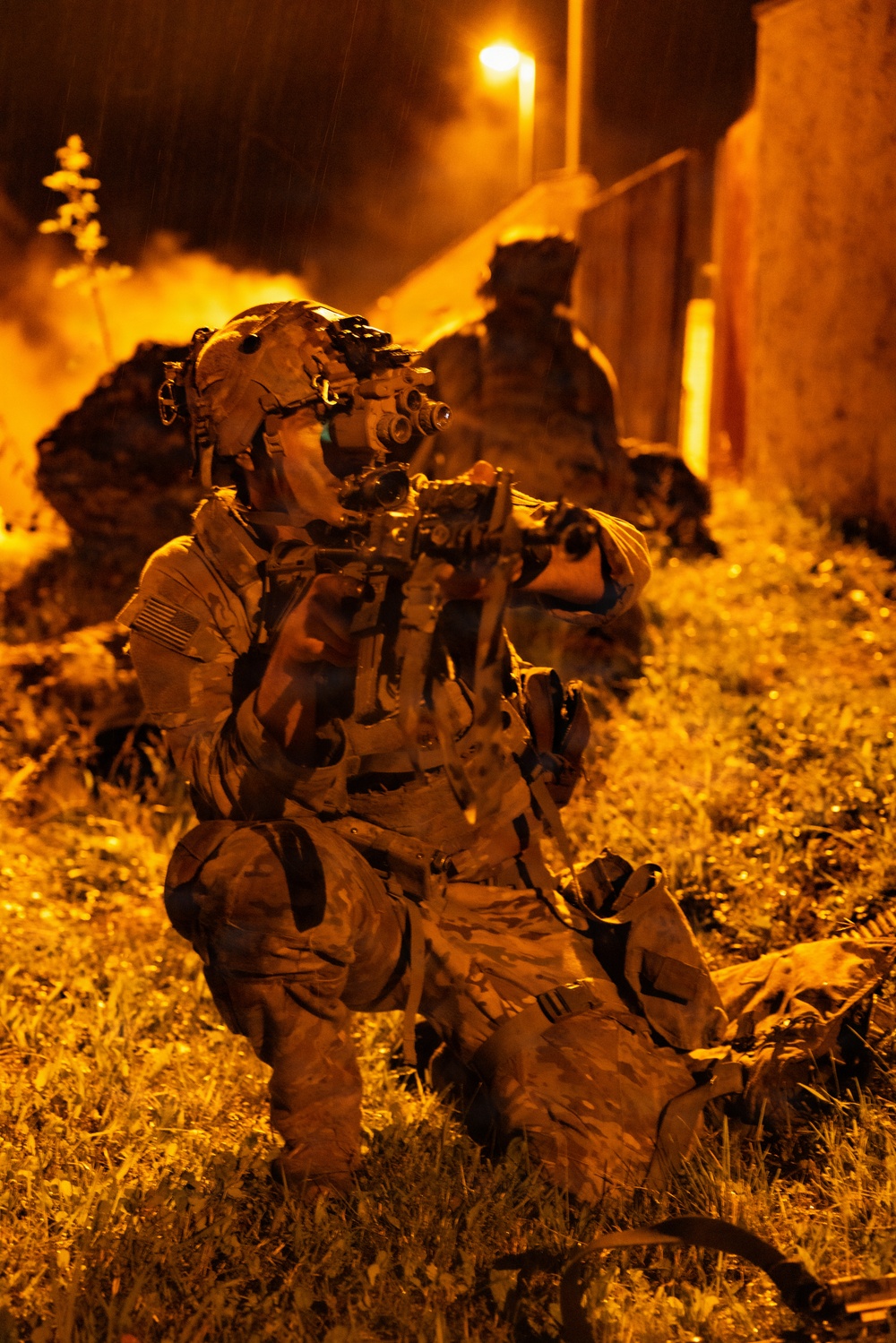 A U.S. military special operations soldier is positioned in a kneeling stance during a nighttime operation, aiming a rifle equipped with optics while wearing night vision goggles (NVG) mounted on a helmet. The scene is illuminated by orange ambient light, likely from fire or pyrotechnics in the background, with rain visible falling. At least one additional soldier is visible in the background near a structure.