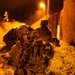 A U.S. military special operations soldier is positioned in a kneeling stance during a nighttime operation, aiming a rifle equipped with optics while wearing night vision goggles (NVG) mounted on a helmet. The scene is illuminated by orange ambient light, likely from fire or pyrotechnics in the background, with rain visible falling. At least one additional soldier is visible in the background near a structure.