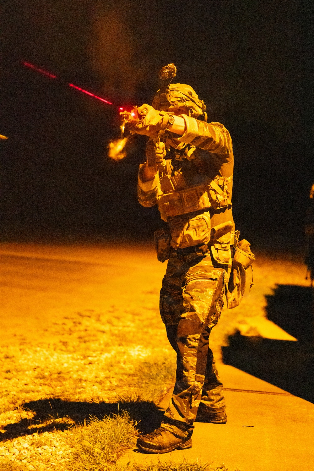 A heavily equipped soldier or operator is photographed at night, actively firing a rifle with a visible red laser sight beam projecting forward. The subject is wearing full tactical gear including a plate carrier, helmet with mounted optics (likely night vision device), camouflage uniform, and multiple pouches. The scene is illuminated by ambient orange/sodium lighting from below, creating a dramatic contrast against the dark background.