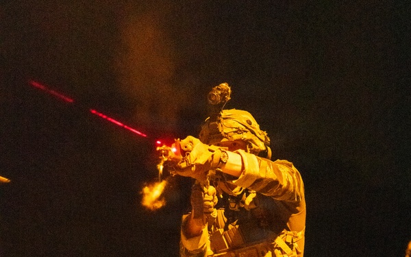 A heavily equipped soldier or operator is photographed at night, actively firing a rifle with a visible red laser sight beam projecting forward. The subject is wearing full tactical gear including a plate carrier, helmet with mounted optics (likely night vision device), camouflage uniform, and multiple pouches. The scene is illuminated by ambient orange/sodium lighting from below, creating a dramatic contrast against the dark background.