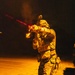 A heavily equipped soldier or operator is photographed at night, actively firing a rifle with a visible red laser sight beam projecting forward. The subject is wearing full tactical gear including a plate carrier, helmet with mounted optics (likely night vision device), camouflage uniform, and multiple pouches. The scene is illuminated by ambient orange/sodium lighting from below, creating a dramatic contrast against the dark background.