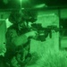 A night-vision captured image showing armed military personnel conducting an operation in what appears to be an urban or compound environment. The primary subject in the foreground is aiming a rifle equipped with tactical accessories, while a second soldier is visible in the background. The characteristic green monochrome tones confirm this was recorded through night-vision optics.