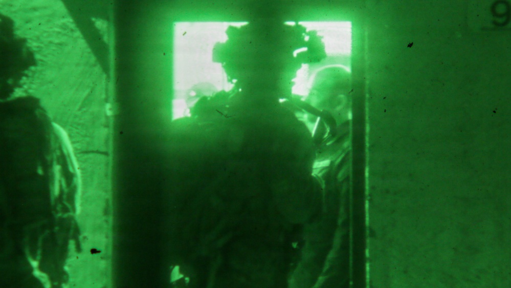 A night-vision (NVG) captured image showing multiple armed personnel in tactical gear conducting what appears to be a building entry or room-clearing operation. The green monochromatic imagery is characteristic of Generation 2 or 3 night-vision equipment. Personnel are positioned at or near a doorway or window opening with bright light emanating from beyond.