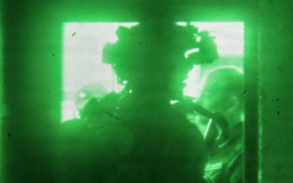 A night-vision (NVG) captured image showing multiple armed personnel in tactical gear conducting what appears to be a building entry or room-clearing operation. The green monochromatic imagery is characteristic of Generation 2 or 3 night-vision equipment. Personnel are positioned at or near a doorway or window opening with bright light emanating from beyond.