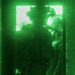 A night-vision (NVG) captured image showing multiple armed personnel in tactical gear conducting what appears to be a building entry or room-clearing operation. The green monochromatic imagery is characteristic of Generation 2 or 3 night-vision equipment. Personnel are positioned at or near a doorway or window opening with bright light emanating from beyond.