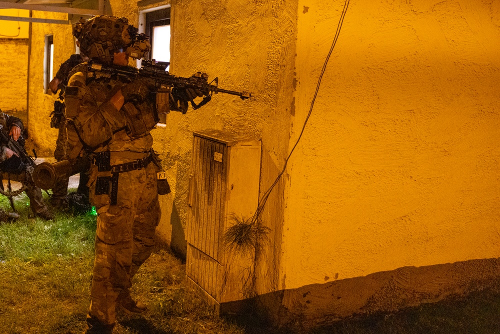 A heavily equipped soldier in full combat gear is positioned against a yellow stucco building wall at night, aiming a rifle around a corner. The scene is illuminated by artificial orange/yellow lighting, suggesting a nighttime military training exercise or operation. At least one additional armed personnel is visible in the background to the left.