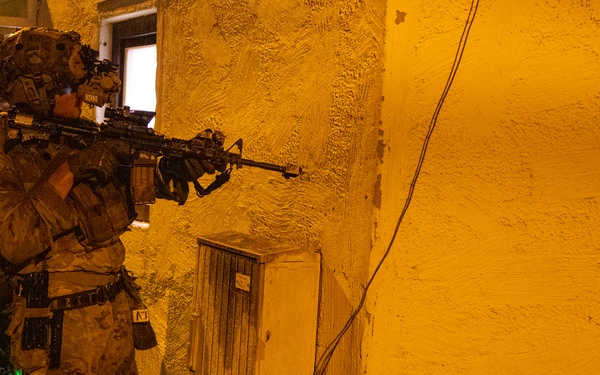 A heavily equipped soldier in full combat gear is positioned against a yellow stucco building wall at night, aiming a rifle around a corner. The scene is illuminated by artificial orange/yellow lighting, suggesting a nighttime military training exercise or operation. At least one additional armed personnel is visible in the background to the left.