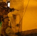 A heavily equipped soldier in full combat gear is positioned against a yellow stucco building wall at night, aiming a rifle around a corner. The scene is illuminated by artificial orange/yellow lighting, suggesting a nighttime military training exercise or operation. At least one additional armed personnel is visible in the background to the left.