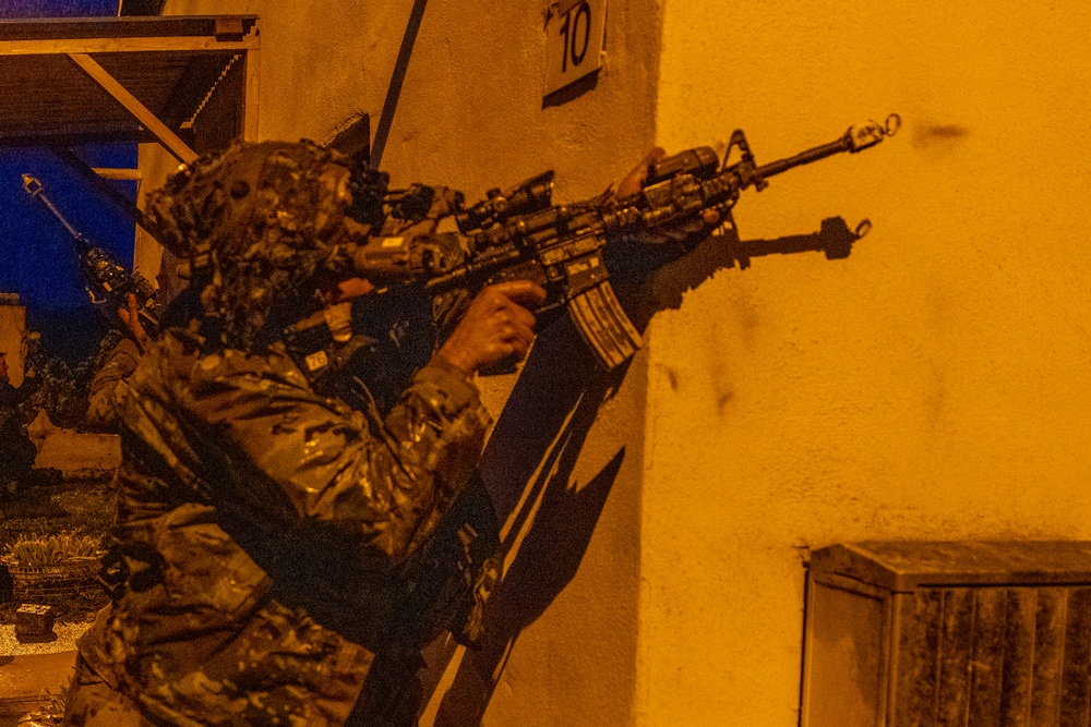 A heavily equipped soldier in camouflage uniform is aiming a rifle with multiple attachments around the corner of a building at night. The scene is illuminated by orange artificial lighting, suggesting an urban nighttime operation. At least one additional armed individual is visible in the background.