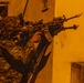 A heavily equipped soldier in camouflage uniform is aiming a rifle with multiple attachments around the corner of a building at night. The scene is illuminated by orange artificial lighting, suggesting an urban nighttime operation. At least one additional armed individual is visible in the background.