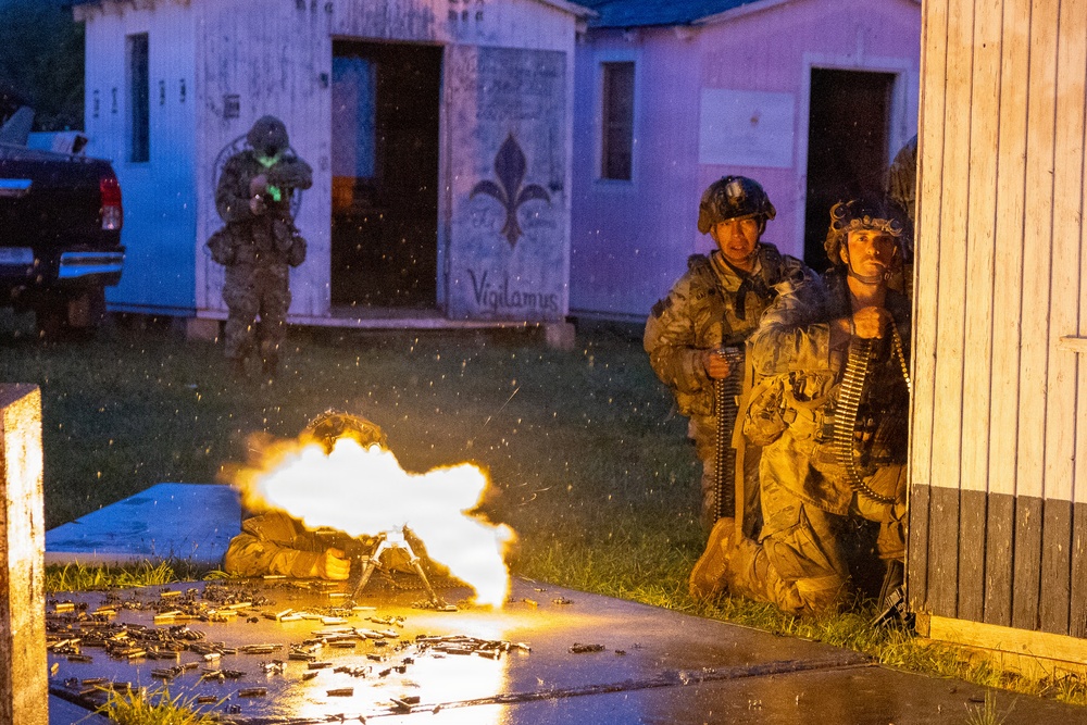 A nighttime military training exercise showing soldiers in tactical positions around a simulated urban environment. A machine gun is firing with a visible muzzle flash and spent brass casings scattered on the ground. Multiple soldiers are visible in different positions, with the word 'Vigilamus' (Latin for 'We Watch') painted on a building in the background.