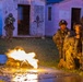 A nighttime military training exercise showing soldiers in tactical positions around a simulated urban environment. A machine gun is firing with a visible muzzle flash and spent brass casings scattered on the ground. Multiple soldiers are visible in different positions, with the word 'Vigilamus' (Latin for 'We Watch') painted on a building in the background.