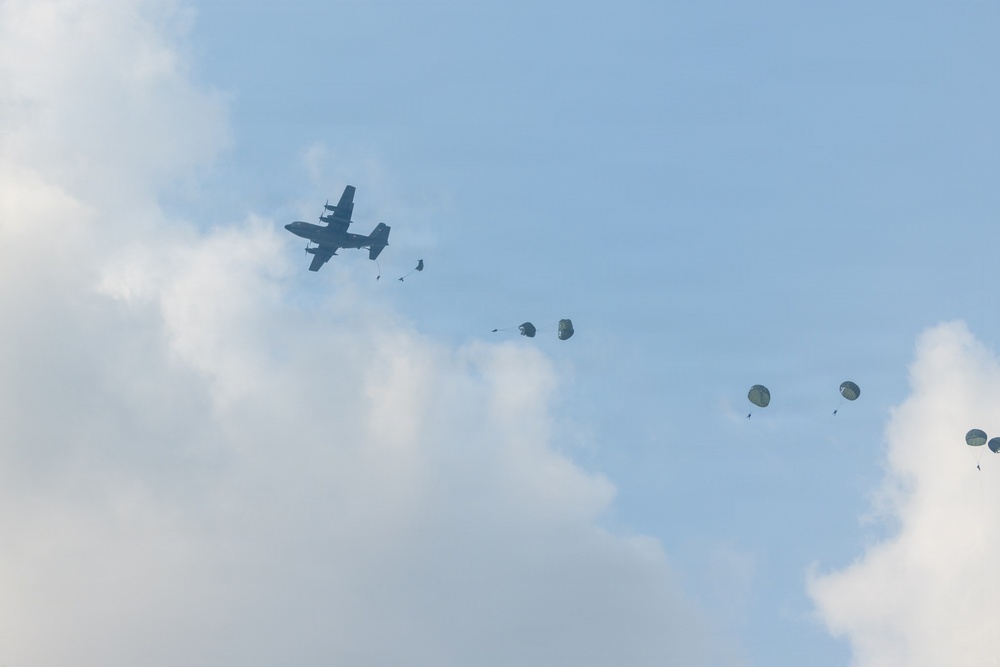 A military transport aircraft, consistent with a C-130 Hercules silhouette, is conducting an airborne drop operation against a partly cloudy sky. Multiple paratroopers or cargo bundles can be seen at various stages of deployment, with parachutes in different states of inflation trailing behind the aircraft. The image captures a sequential mass-exit jump with approximately 4-5 deployed parachutes visible.