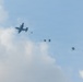 A military transport aircraft, consistent with a C-130 Hercules silhouette, is conducting an airborne drop operation against a partly cloudy sky. Multiple paratroopers or cargo bundles can be seen at various stages of deployment, with parachutes in different states of inflation trailing behind the aircraft. The image captures a sequential mass-exit jump with approximately 4-5 deployed parachutes visible.