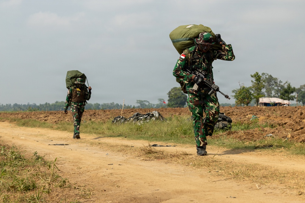 Two soldiers in camouflage uniforms are walking along a dirt path in an open field environment, carrying large military rucksacks on their backs. The foreground soldier is also carrying an assault rifle and displays a red-and-white flag patch on their shoulder, consistent with the Indonesian flag. The setting appears to be a military training or operational area with tilled fields and sparse vegetation in the background.