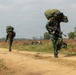 Two soldiers in camouflage uniforms are walking along a dirt path in an open field environment, carrying large military rucksacks on their backs. The foreground soldier is also carrying an assault rifle and displays a red-and-white flag patch on their shoulder, consistent with the Indonesian flag. The setting appears to be a military training or operational area with tilled fields and sparse vegetation in the background.