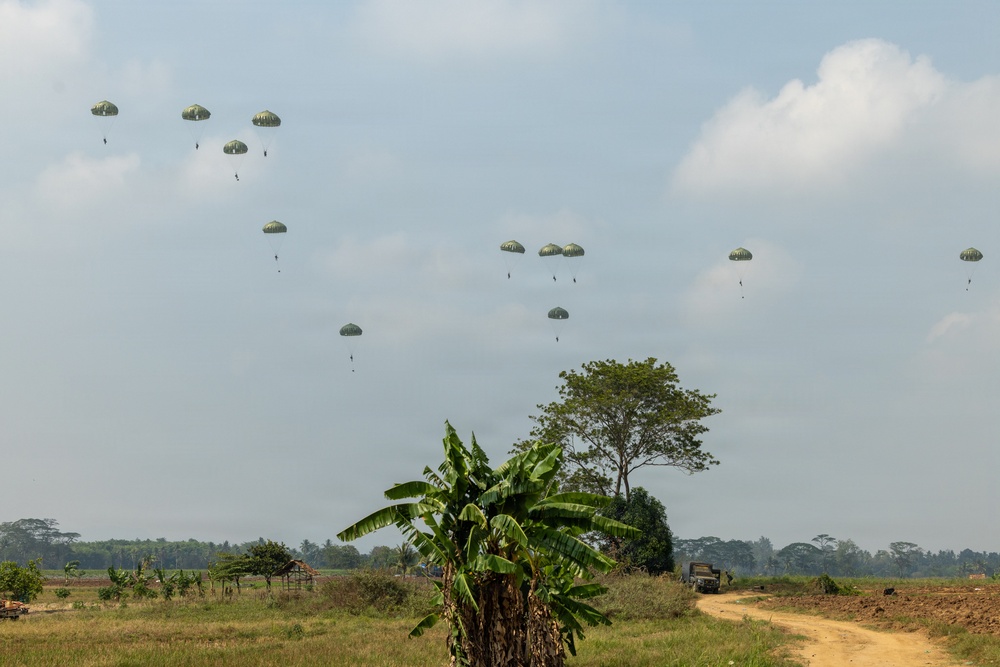 A military airborne operation is underway, with numerous paratroopers descending from the sky using olive-green/camouflage parachutes over a rural, tropical landscape. The drop zone appears to be an open field with scattered vegetation including banana plants and trees. A dirt road and a military vehicle are visible in the foreground, suggesting a coordinated tactical exercise or operation.