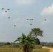 A military airborne operation is underway, with numerous paratroopers descending from the sky using olive-green/camouflage parachutes over a rural, tropical landscape. The drop zone appears to be an open field with scattered vegetation including banana plants and trees. A dirt road and a military vehicle are visible in the foreground, suggesting a coordinated tactical exercise or operation.