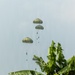 The image shows multiple military paratroopers descending with olive-green round parachutes over a tropical rural landscape. At least four parachutes with suspended figures are visible at varying altitudes, suggesting a sequential jump from an aircraft. The setting features banana trees in the foreground, a small thatched shelter, open grassland, and dense tropical vegetation in the background.