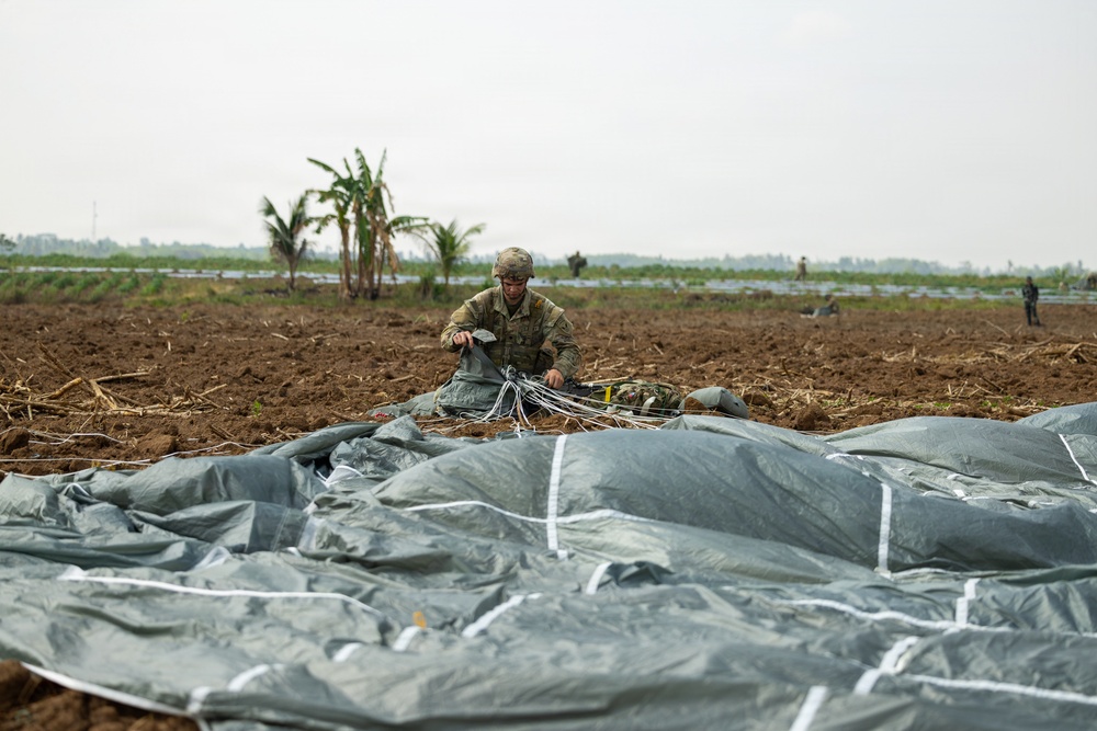 A soldier in camouflage uniform is kneeling in a plowed agricultural field, gathering and packing parachute lines and canopy after a landing. A large gray parachute canopy is spread across the foreground of the image. Additional personnel are visible in the background of the open field, which appears to be in a tropical region based on the banana trees visible.