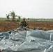A soldier in camouflage uniform is kneeling in a plowed agricultural field, gathering and packing parachute lines and canopy after a landing. A large gray parachute canopy is spread across the foreground of the image. Additional personnel are visible in the background of the open field, which appears to be in a tropical region based on the banana trees visible.