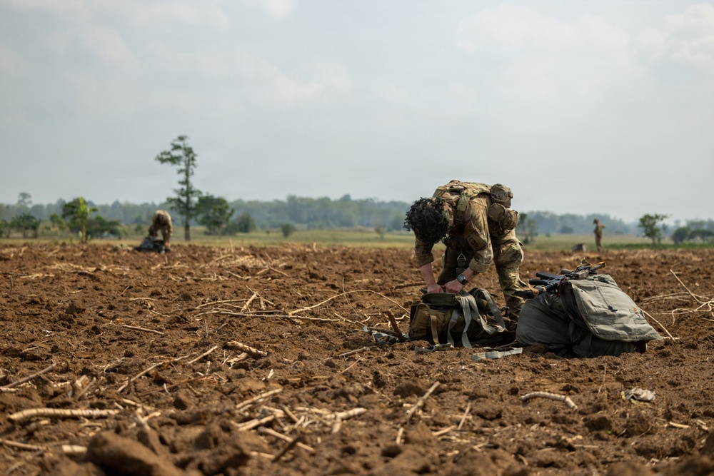 Military personnel in camouflage uniforms are dispersed across a plowed agricultural field, conducting what appears to be a field exercise or post-landing operation. The foreground shows one soldier bending over military gear bags, while additional soldiers are visible in the background at varying distances. The terrain is dry, tilled soil with crop stubble remnants, set against a hazy treeline horizon.