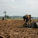 Military personnel in camouflage uniforms are dispersed across a plowed agricultural field, conducting what appears to be a field exercise or post-landing operation. The foreground shows one soldier bending over military gear bags, while additional soldiers are visible in the background at varying distances. The terrain is dry, tilled soil with crop stubble remnants, set against a hazy treeline horizon.