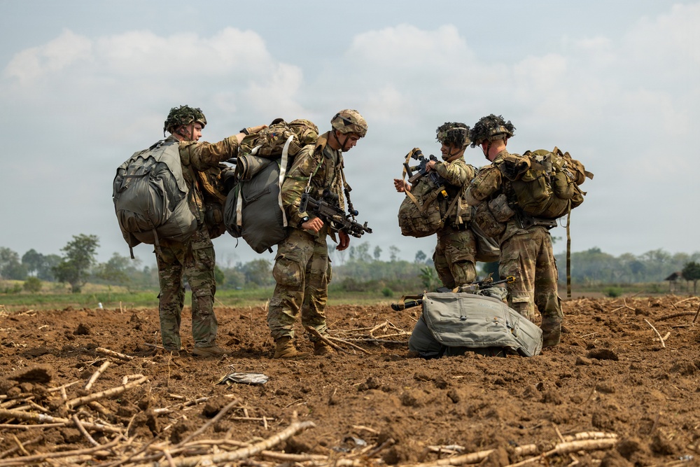 Four soldiers in multicam/OCP camouflage uniforms are gathered in an open dirt field, appearing to consolidate and check equipment after what appears to be a parachute landing operation. They are carrying large parachute deployment bags and tactical gear including weapons. The setting appears to be a rural agricultural area with flat terrain and scattered trees in the background.