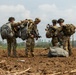 Four soldiers in multicam/OCP camouflage uniforms are gathered in an open dirt field, appearing to consolidate and check equipment after what appears to be a parachute landing operation. They are carrying large parachute deployment bags and tactical gear including weapons. The setting appears to be a rural agricultural area with flat terrain and scattered trees in the background.
