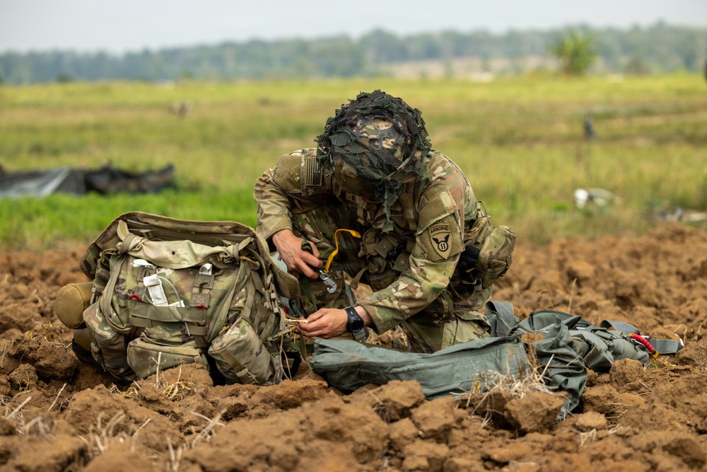 A soldier wearing multicam camouflage uniform and a ghillie-style face covering is kneeling on disturbed dirt terrain, handling equipment after what appears to be a parachute landing. The soldier has a unit patch visible on the left shoulder, identifiable as the 11th Airborne Division patch. A military rucksack is positioned to the left, and parachute rigging or canopy material is spread to the right.