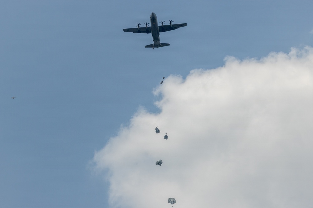 A military transport aircraft is conducting an airborne operation, with multiple paratroopers visible at various stages of descent below the aircraft. The paratroopers' parachutes appear to be in different phases of deployment, from just exiting the aircraft to partially or fully opened canopies. The scene is set against a partly cloudy blue sky.