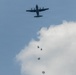 A military transport aircraft is conducting an airborne operation, with multiple paratroopers visible at various stages of descent below the aircraft. The paratroopers' parachutes appear to be in different phases of deployment, from just exiting the aircraft to partially or fully opened canopies. The scene is set against a partly cloudy blue sky.