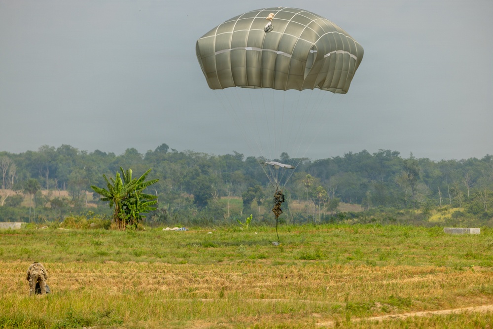 A military parachute operation is underway over an open grassy field, with at least two parachutists visible at different stages of descent. One large olive/grey military parachute is prominently deployed in the upper portion of the image, while a second smaller parachute with a suspended figure is visible in the mid-ground. A third individual in military gear is visible on the ground in the lower left, having already landed.