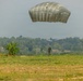 A military parachute operation is underway over an open grassy field, with at least two parachutists visible at different stages of descent. One large olive/grey military parachute is prominently deployed in the upper portion of the image, while a second smaller parachute with a suspended figure is visible in the mid-ground. A third individual in military gear is visible on the ground in the lower left, having already landed.
