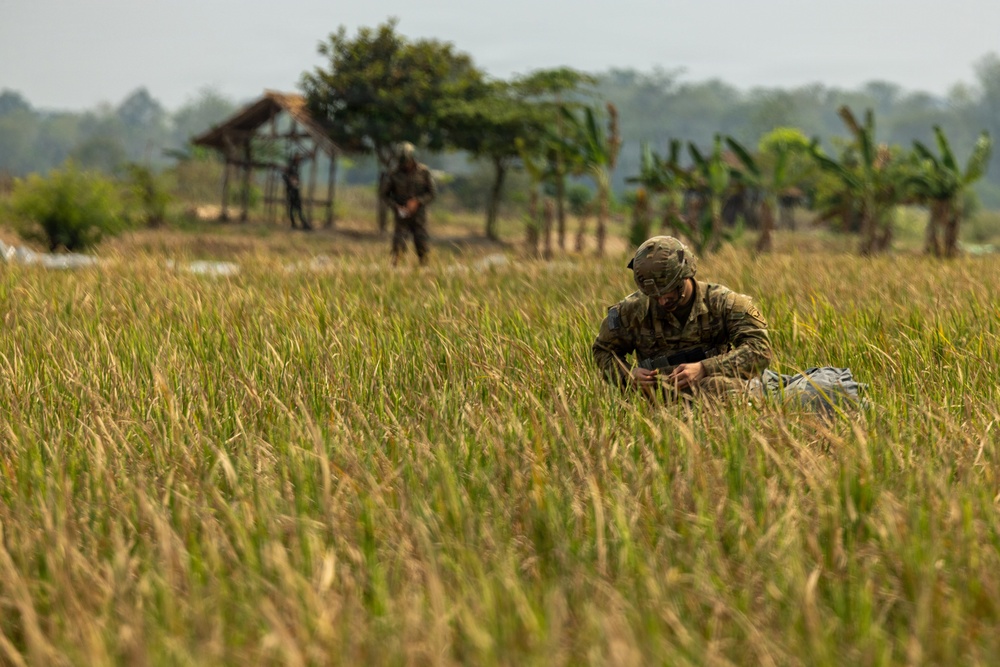 A soldier in camouflage uniform and helmet is crouched low in a tall grass field, appearing to handle equipment or a pack. Two additional soldiers are visible in the background near a rustic wooden structure, suggesting a military field exercise or patrol operation. The setting appears to be a rural agricultural area in a tropical region, indicated by banana trees and lush vegetation.