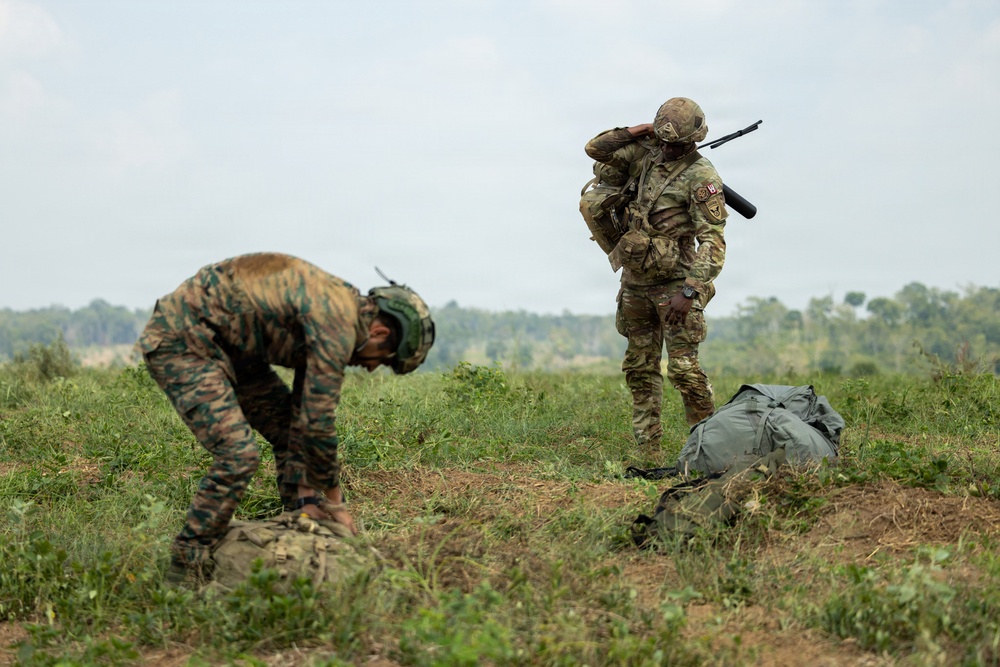 Two military personnel are visible in an open grassy field with treeline in the background. One soldier wearing tiger-stripe camouflage and a green helmet is bent over attending to gear on the ground, while a second soldier in multicam/OCP pattern uniform stands upright with a rifle slung over their shoulder. Both have tactical equipment and appear to be in a post-landing or field assembly scenario.