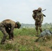 Two military personnel are visible in an open grassy field with treeline in the background. One soldier wearing tiger-stripe camouflage and a green helmet is bent over attending to gear on the ground, while a second soldier in multicam/OCP pattern uniform stands upright with a rifle slung over their shoulder. Both have tactical equipment and appear to be in a post-landing or field assembly scenario.