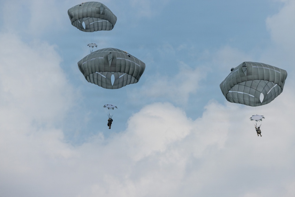 The image shows multiple military paratroopers descending from the sky using round, olive-drab/gray military parachutes. At least five parachutes are visible at varying altitudes against a partly cloudy blue sky. The parachutes appear to be standard military static-line round canopies, consistent with airborne operations.